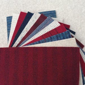 May include: A fan of various fabric swatches in shades of red, blue, white, and beige. The swatches are arranged in a semi-circle, showcasing different textures and patterns, including herringbone and woven designs. The color palette is a mix of warm and cool tones.