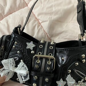 Puede incluir: Bolso de mano negro con herrajes plateados, incluyendo cremalleras, tachuelas y adornos en forma de estrella. El bolso presenta una correa con tachuelas y un lazo de encaje blanco. Acabado brillante y dise&ntilde;o estructurado.