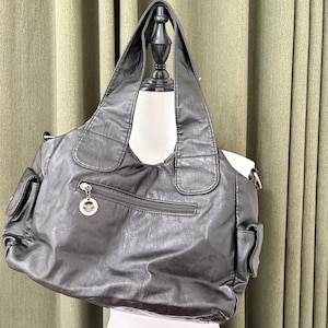 May include: A black faux leather tote bag with two exterior pockets and a zippered main compartment. The bag has two shoulder straps and a silver-tone charm. The bag is displayed on a white mannequin.