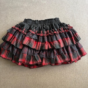 May include: A black and red plaid mini skirt with multiple ruffled layers. The skirt features black lace accents between the plaid ruffles and a black elastic waistband. The skirt is a stylish and unique piece of clothing.