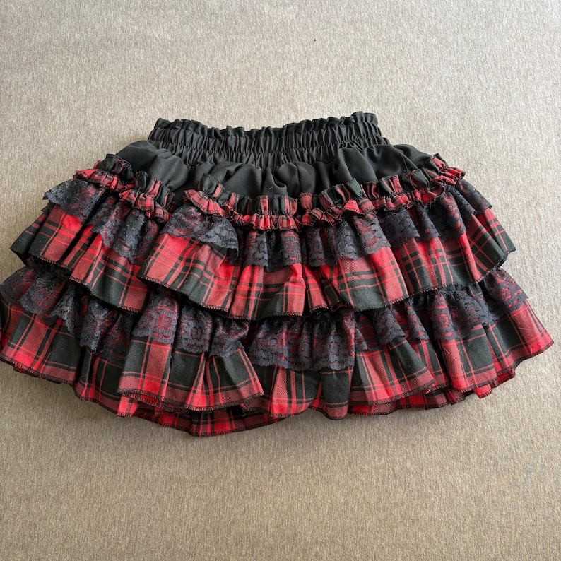May include: A black and red plaid mini skirt with multiple ruffled layers. The skirt features black lace accents between the plaid ruffles and a black elastic waistband. The skirt is a stylish and unique piece of clothing.
