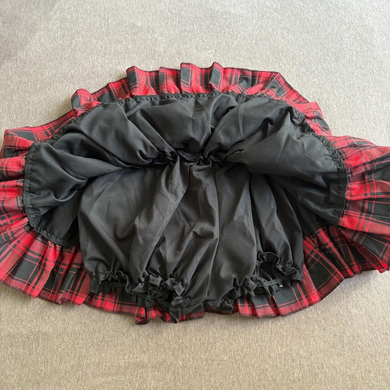 May include: A red and black plaid skirt with a black lining. The skirt is pleated and has a ruffled hem. The skirt is made of a lightweight fabric.