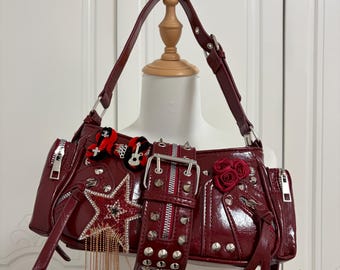 May include: A burgundy shoulder bag with a glossy finish, adorned with a star, studs, and a decorative belt. Features include a zipper closure, a guitar-shaped embellishment, and a rose accent. The bag has a shoulder strap.