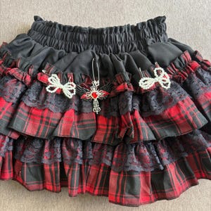 May include: Black and red plaid skirt with multiple ruffled layers. The skirt features black lace trim and is adorned with decorative pins, including a cross pendant with a red gem and white bow accents. The skirt has an elastic waistband.