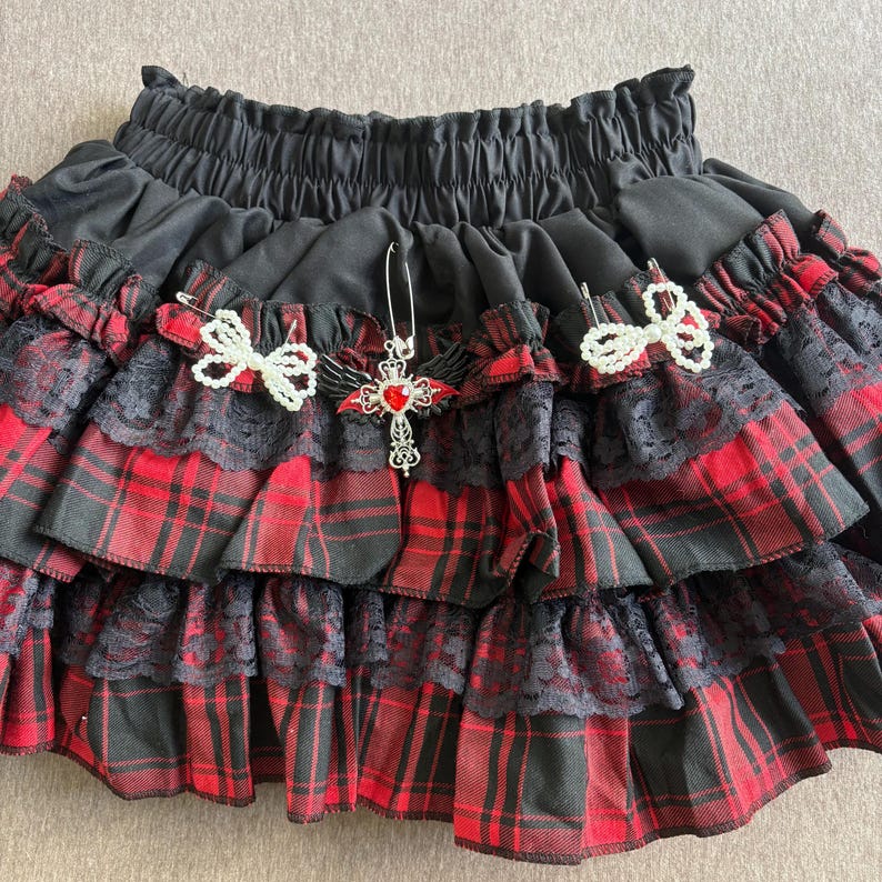 May include: Black and red plaid skirt with multiple ruffled layers. The skirt features black lace trim and is adorned with decorative pins, including a cross pendant with a red gem and white bow accents. The skirt has an elastic waistband.