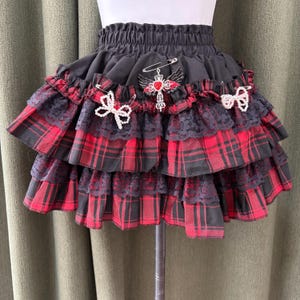 May include: A tiered skirt with a black elastic waistband and layers of red and black plaid fabric. The skirt is adorned with black lace trim, decorative pins, and white bow accents. A cross pendant with red accents is pinned to the skirt.