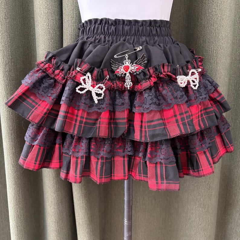 May include: A tiered skirt with a black elastic waistband and layers of red and black plaid fabric. The skirt is adorned with black lace trim, decorative pins, and white bow accents. A cross pendant with red accents is pinned to the skirt.