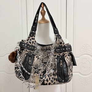 Peut inclure: Sac à bandoulière à imprimé léopard avec des détails noirs, des fermetures éclair et des chaînes argentées. Le sac comporte un pompon en fourrure marron, des étoiles et une chaîne cloutée. Le sac est présenté sur un mannequin.