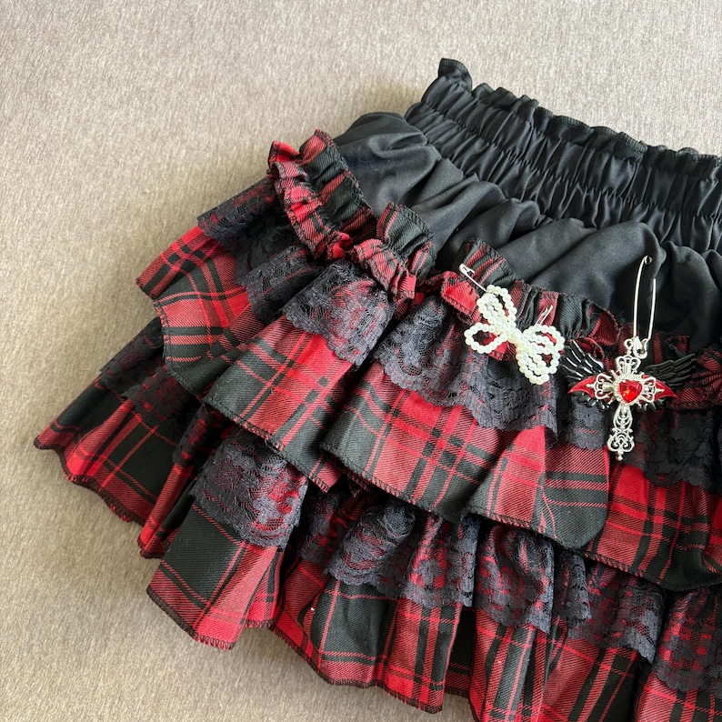 May include: A black and red plaid mini skirt with multiple ruffled layers. Black lace trim accents the plaid fabric. A decorative pin with a red heart and cross is attached to the skirt.