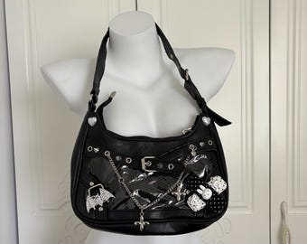 May include: Black shoulder bag with a glossy finish, adorned with silver-tone studs, a buckle, and a chain. Features a bat-shaped charm, a cross pendant, and a black and white polka dot bow with a bunny. The bag has a zipper closure and a heart-shaped detail.