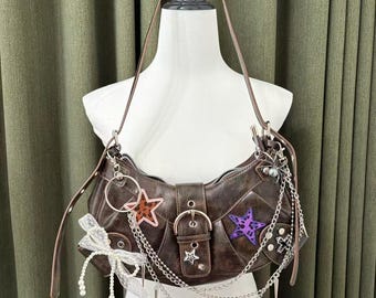 May include: Brown leather shoulder bag with star patches in orange and purple. The bag features silver chain accents, a buckle closure, and a white bow with pearl details. The bag is displayed on a white mannequin.