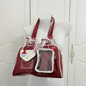 Può includere: Una borsa a mano rosso scuro lucida con bordi bianchi, dotata di due tracolle, una tasca con patta bianca e una finestra rettangolare trasparente. La borsa è decorata con fiocchi in pizzo bianco e una piccola spilla circolare con caratteri.