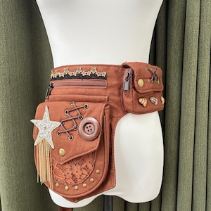 Peut inclure: Sac banane marron de style steampunk avec plusieurs poches et éléments décoratifs. Comprend une étoile en strass avec des franges dorées, de la dentelle et des clous métalliques. Le sac ceinture a une fermeture éclair et un gros bouton en bois.