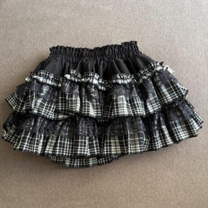 May include: A black and white plaid skirt with multiple ruffled layers. The skirt has a black elastic waistband and is made of a plaid fabric with lace accents. The skirt is designed with a tiered, layered style.