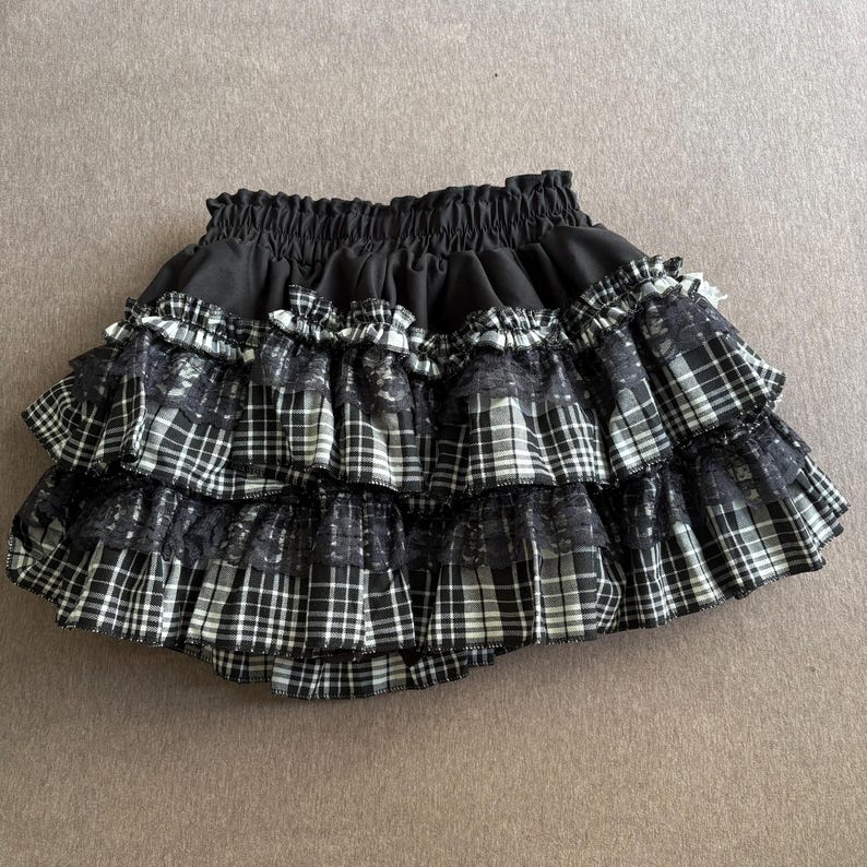 May include: A black and white plaid skirt with multiple ruffled layers. The skirt has a black elastic waistband and is made of a plaid fabric with lace accents. The skirt is designed with a tiered, layered style.