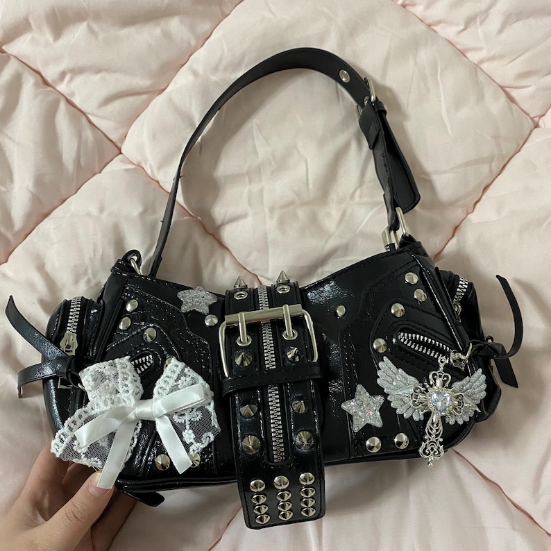 Puede incluir: Bolso de mano negro con tachuelas plateadas, cremalleras y cierre de hebilla. Cuenta con un lazo de encaje blanco, dijes de estrella y cruz, y un asa superior. El bolso tiene un acabado brillante y un cintur&oacute;n con tachuelas.