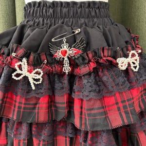 May include: A black and red plaid skirt with multiple layers of ruffles and lace trim. The skirt is adorned with decorative pins, including a cross with wings and a heart, and two pearl bow pins. The skirt has an elastic waistband.