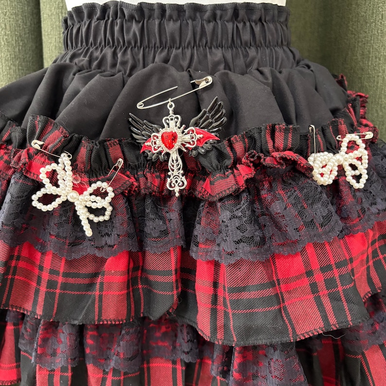 May include: A black and red plaid skirt with multiple layers of ruffles and lace trim. The skirt is adorned with decorative pins, including a cross with wings and a heart, and two pearl bow pins. The skirt has an elastic waistband.
