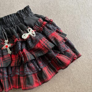 May include: A layered, ruffled skirt in a red and black plaid pattern. The skirt features black lace trim and is adorned with decorative pins, including a cross pendant with a red gem and a white bow. The waistband is black and elasticized.