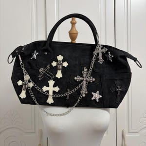 Borsa a tracolla in pelle scamosciata nera in stile gotico rock con applicazioni a croce, catena argentata, borchie a stella e spille da balia, ideale per un look alternativo anni 2000.