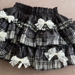 May include: Black and white plaid skirt with multiple ruffled layers. The skirt features white lace bows and a black elastic waistband. The skirt has a gothic or Lolita aesthetic.