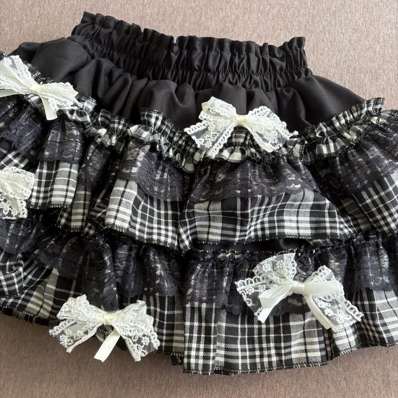 May include: Black and white plaid skirt with multiple ruffled layers. The skirt features white lace bows and a black elastic waistband. The skirt has a gothic or Lolita aesthetic.