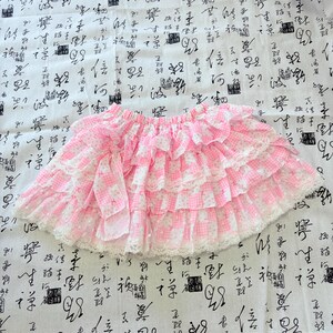 May include: A pink and white gingham skirt featuring multiple horizontal ruffled tiers. The skirt is adorned with delicate white lace trim along the edges of each ruffle and includes a decorative fabric bow on the left side of the waistband.