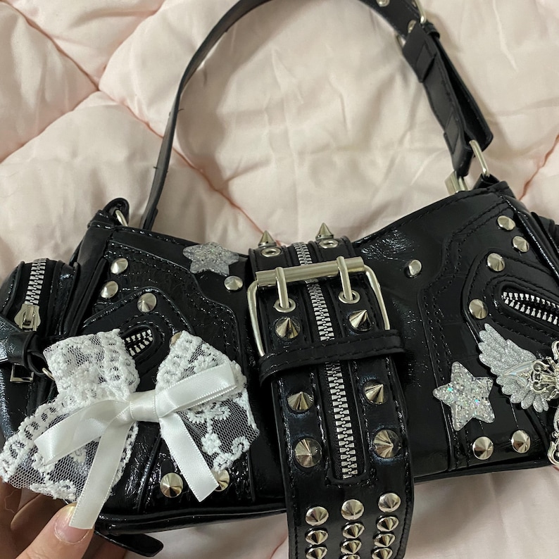 Puede incluir: Bolso de mano negro con tachuelas plateadas, cremalleras y un cintur&oacute;n decorativo. El bolso presenta un lazo de encaje blanco y adornos de estrellas plateadas. La correa es negra y el bolso tiene un acabado brillante.