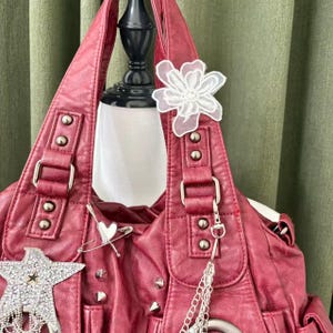 May include: A burgundy faux leather handbag with silver hardware, studs, and decorative charms. The bag features a white lace flower, a star-shaped charm, and a safety pin with a heart. The bag is displayed on a mannequin.