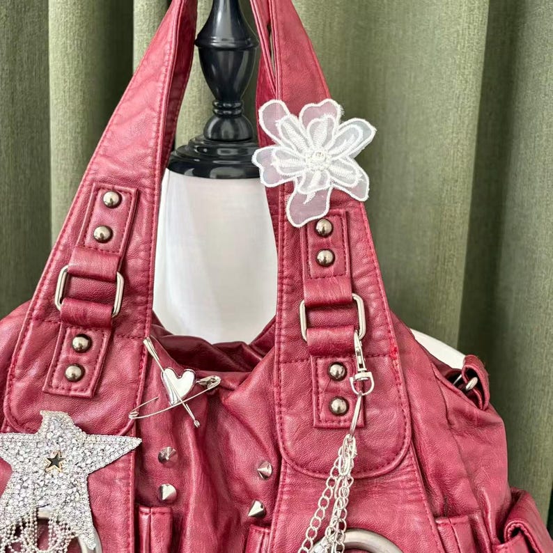 May include: A burgundy faux leather handbag with silver hardware, studs, and decorative charms. The bag features a white lace flower, a star-shaped charm, and a safety pin with a heart. The bag is displayed on a mannequin.