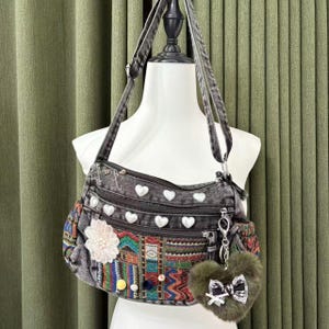 Peut inclure: Un sac bandoulière en jean gris avec plusieurs compartiments zippés et des ornements blancs en forme de cœur. Le sac présente un panneau brodé coloré et un porte-clés en forme de cœur vert duveteux avec un nœud. Le sac est présenté sur un mannequin.