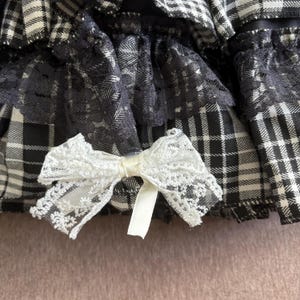 May include: Close-up of a black and white plaid fabric with a white lace bow. The fabric has a layered design with black lace trim. The bow is made of delicate white lace and has a satin ribbon center.