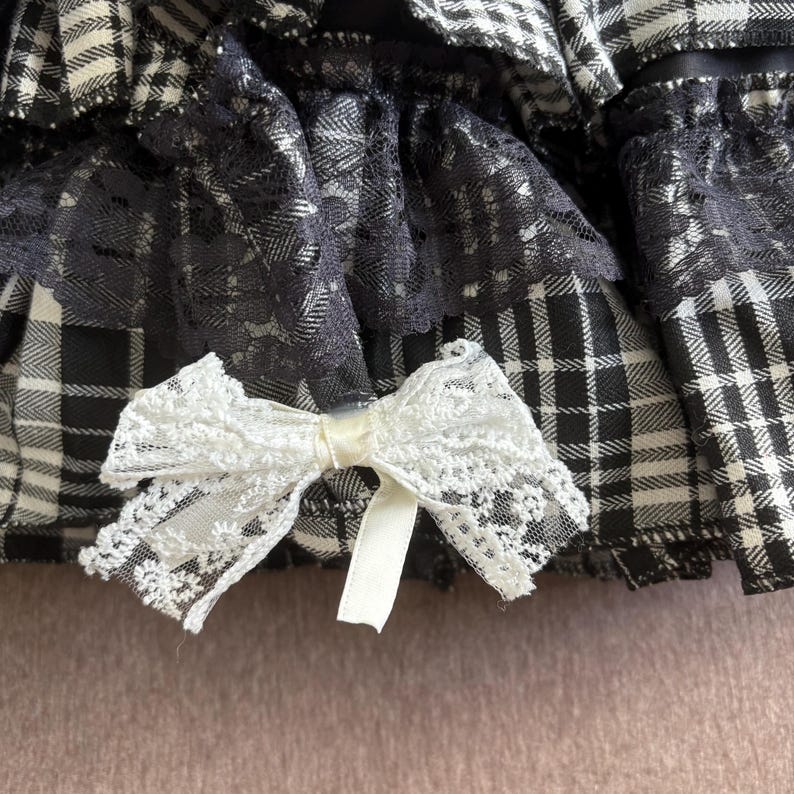May include: Close-up of a black and white plaid fabric with a white lace bow. The fabric has a layered design with black lace trim. The bow is made of delicate white lace and has a satin ribbon center.