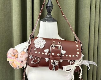 May include: A brown leather shoulder bag with silver studs, hearts, and a flower embellishment. The bag features a buckle closure with a cross pendant and a detachable keychain with a fluffy charm. A white ribbon with pearls is attached.
