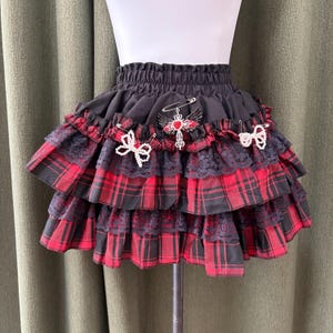 May include: Black and red plaid mini skirt with multiple ruffled layers. The skirt features black lace trim and is adorned with decorative pins, including a cross and bow accents. The skirt is displayed on a mannequin.