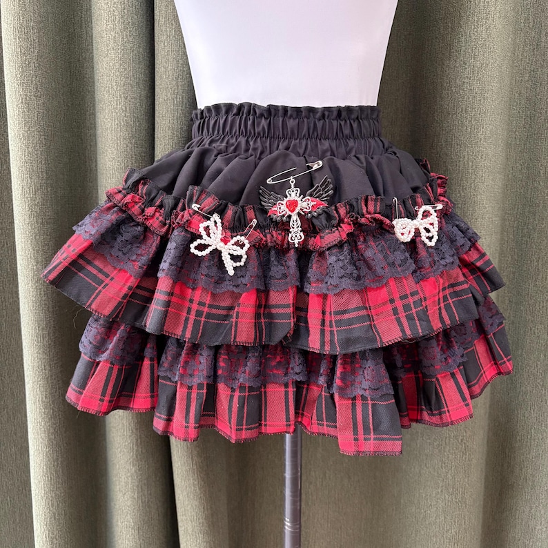 May include: Black and red plaid mini skirt with multiple ruffled layers. The skirt features black lace trim and is adorned with decorative pins, including a cross and bow accents. The skirt is displayed on a mannequin.