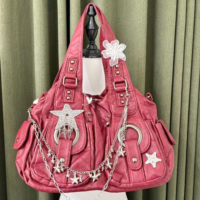 May include: A red handbag with silver star and chain embellishments. The bag has a large, slouchy shape with multiple pockets and a decorative white flower. The bag is displayed on a mannequin.
