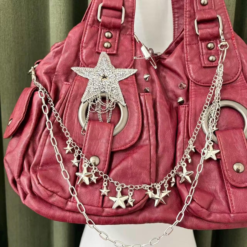 May include: A burgundy handbag with a star-shaped rhinestone embellishment and silver chain details. The bag features multiple pockets, silver hardware, and a chain with star charms. The bag has a textured appearance.