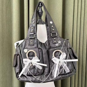 May include: Black faux leather tote bag with multiple pockets, silver chain accents, and white lace bows. The bag features silver hardware, including buckles, rings, and star-shaped embellishments. The bag is displayed on a mannequin.
