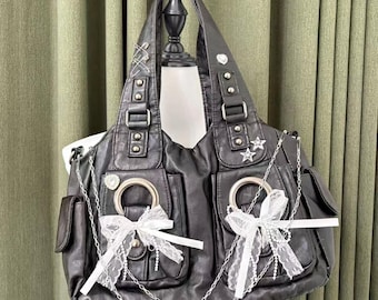 May include: Black faux leather tote bag with multiple pockets, silver chain accents, and white lace bows. The bag features silver hardware, including buckles, rings, and star-shaped embellishments. The bag is displayed on a mannequin.