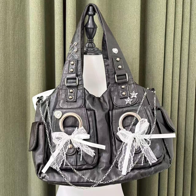 May include: Black faux leather tote bag with multiple pockets, silver chain accents, and white lace bows. The bag features silver hardware, including buckles, rings, and star-shaped embellishments. The bag is displayed on a mannequin.