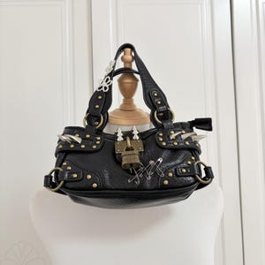 May include: Black handbag with gold-colored studs, spikes, and hardware. The bag features a lock closure and decorative safety pins. A small floral embellishment adorns one of the handles.