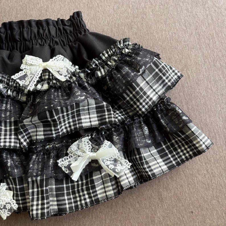 May include: Black and white plaid skirt with lace trim and white bows. The skirt has multiple ruffled layers and a gathered waistband. The plaid pattern is a classic design, and the bows add a delicate touch.