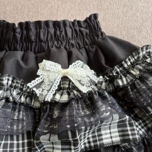 May include: Close-up of a black skirt with a white lace bow. The skirt features a black and white plaid pattern with lace trim. The waistband is gathered, and the overall design is detailed and textured.
