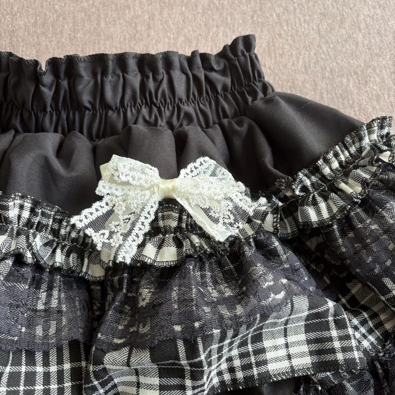 May include: Close-up of a black skirt with a white lace bow. The skirt features a black and white plaid pattern with lace trim. The waistband is gathered, and the overall design is detailed and textured.