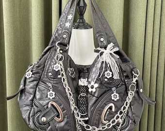 May include: Black faux leather handbag with silver-tone chain and flower charms. The bag features decorative white flower accents, silver rivets, and a lace bow. The bag is displayed on a mannequin.