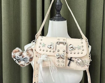 May include: A cream-colored handbag with a shoulder strap, adorned with silver studs, charms, and a buckle closure. The bag features heart, star, and cross embellishments, and a small, fluffy keychain with a bow.