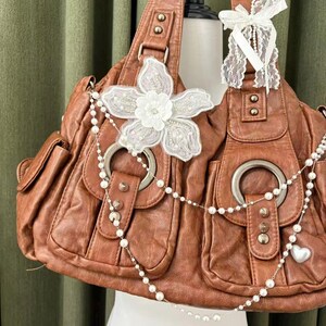 May include: Brown leather handbag with multiple pockets and silver hardware. The bag is decorated with a white floral applique and a pearl necklace. A white lace bow adorns the handle.