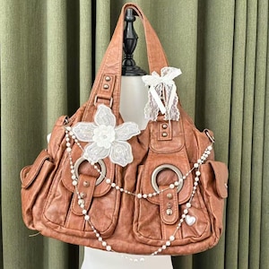 May include: A brown leather handbag with multiple pockets and silver hardware. The bag is decorated with a white lace flower, a pearl chain, and a white lace bow. The bag is displayed on a mannequin.