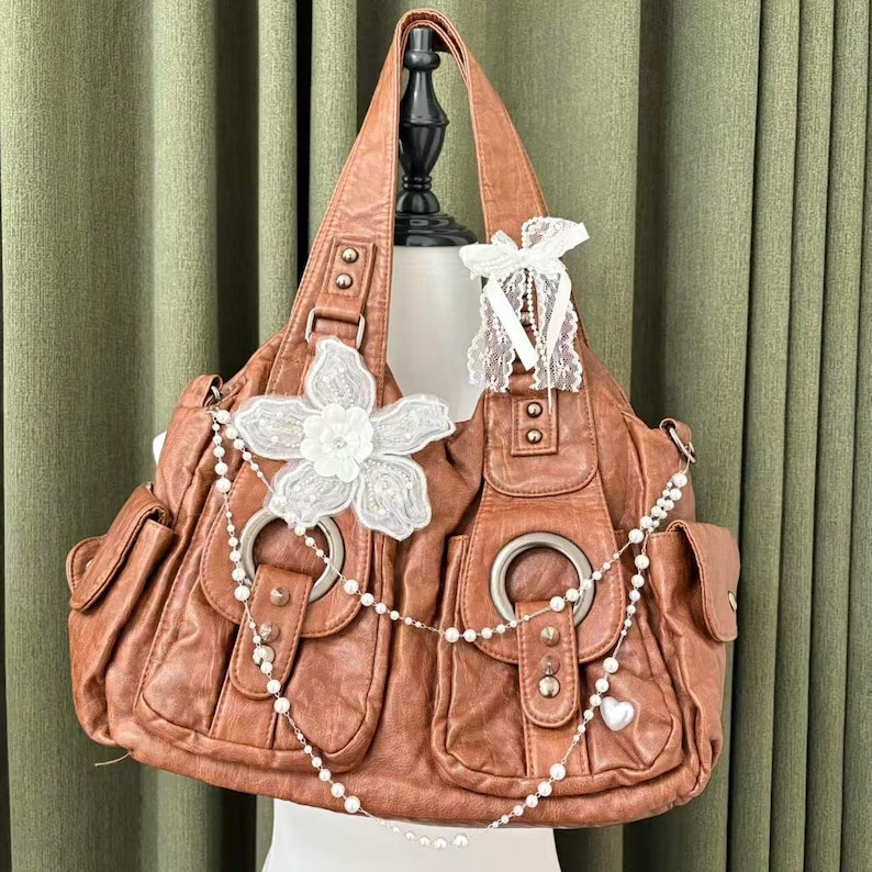 May include: A brown leather handbag with multiple pockets and silver hardware. The bag is decorated with a white lace flower, a pearl chain, and a white lace bow. The bag is displayed on a mannequin.